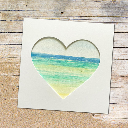 White card with a heart shaped aperture showing a beach, set against a wooden background